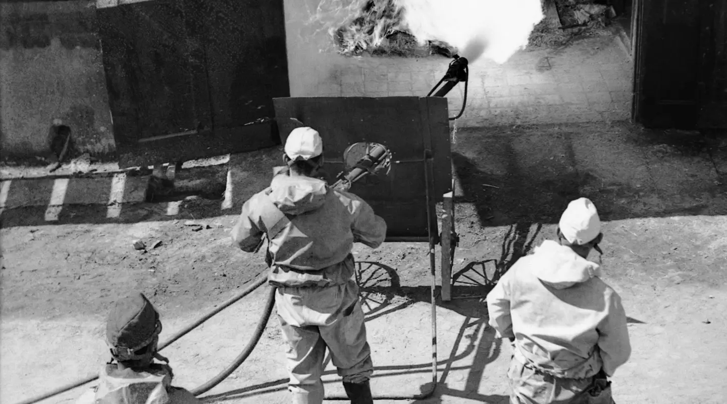 Japanese workmen wearing gas masks directing jet from a mobile flame-thrower towards a pile of gasoline-soaked debris. 
