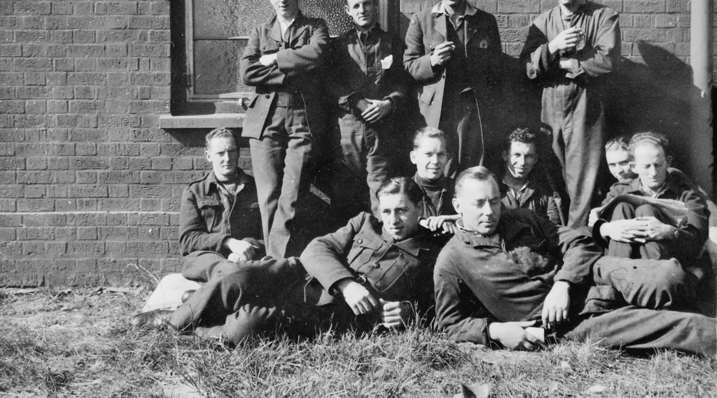 Four men stand against a brick wall, in front are seven men sitting on the ground.