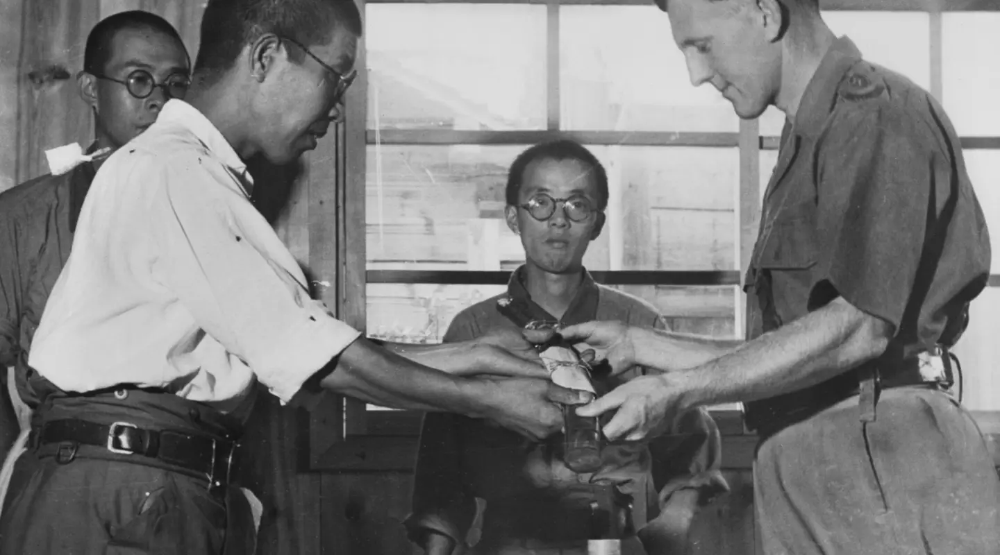 Second Lieutenant Nakanishi formerly Commandant of the Notogawa prisoner of war (POW) camp surrenders his sword (and command) to Lt Goddard who was captured at Singapore on 15 February 1942