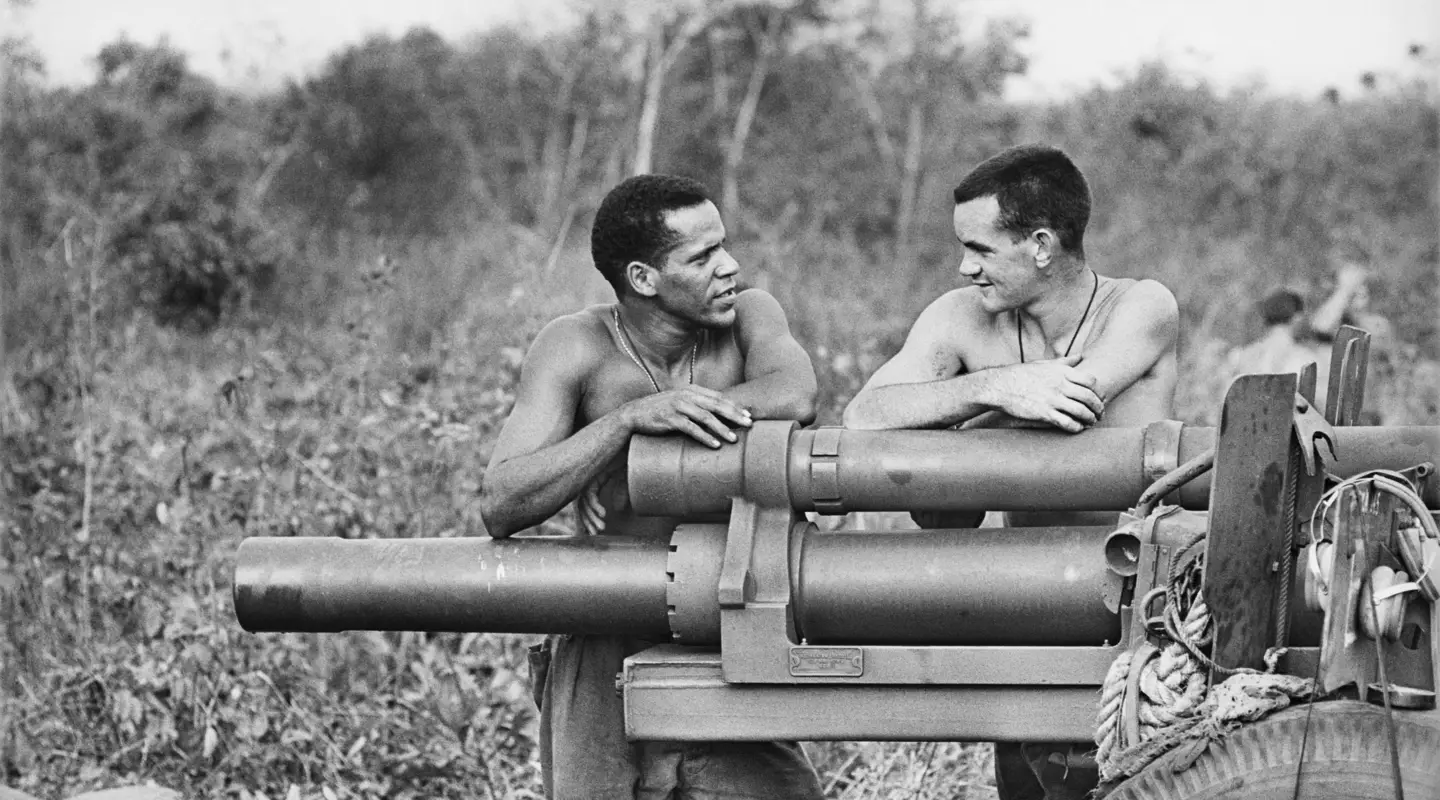 Two men standing, facing each other, arms resting on artillery cannon