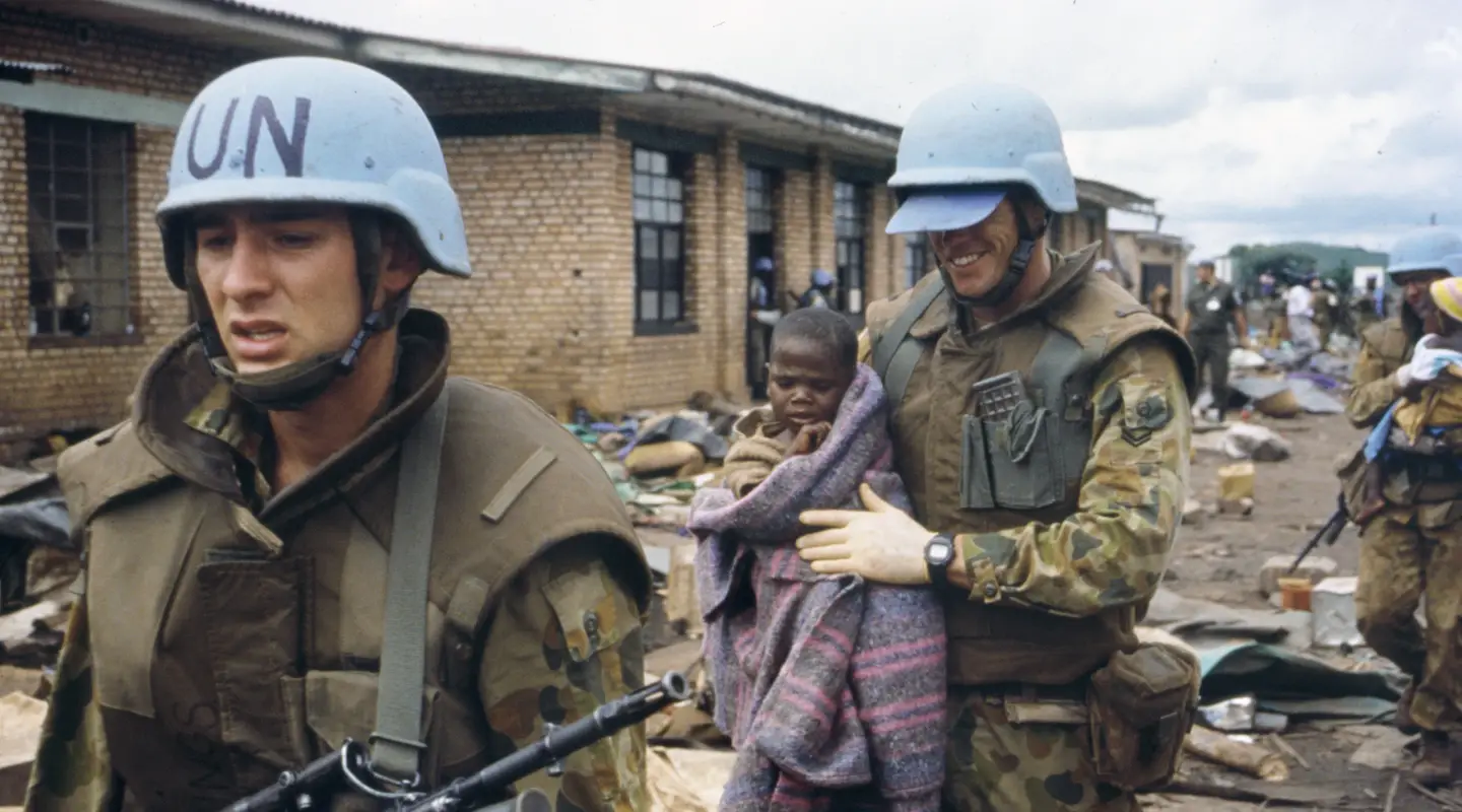 An armed Australian soldier leading another soldier who is carrying a Rwandan child in his arms.