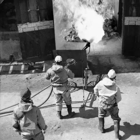 Japanese workmen wearing gas masks directing jet from a mobile flame-thrower towards a pile of gasoline-soaked debris. 