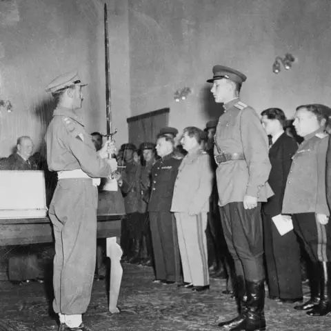 Two people in the foreground, one is holding a long sword in front of this body. Both are in formal uniforms.