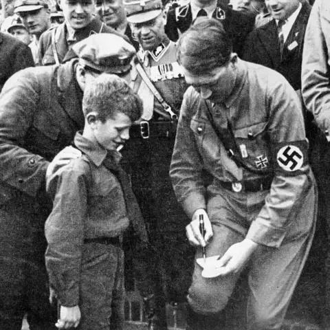 Hitler signs an autograph for a young boy while a crowd looks on