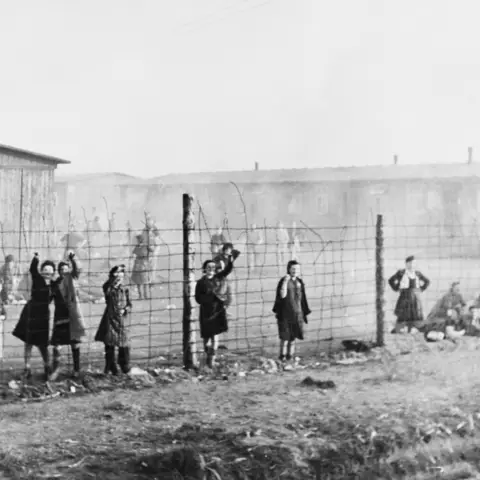 Women standing and waving at the barbed wire fence within Belsen concentration camp.