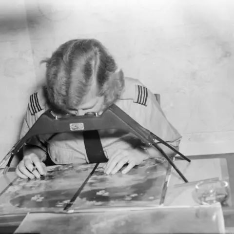 A WAAAF Officer peers through a stereoscope at aerial photographs.