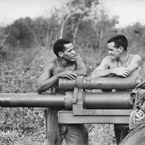 Two men standing, facing each other, arms resting on artillery cannon