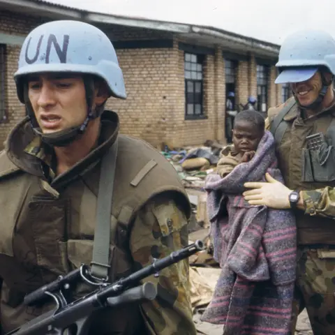 An armed Australian soldier leading another soldier who is carrying a Rwandan child in his arms.