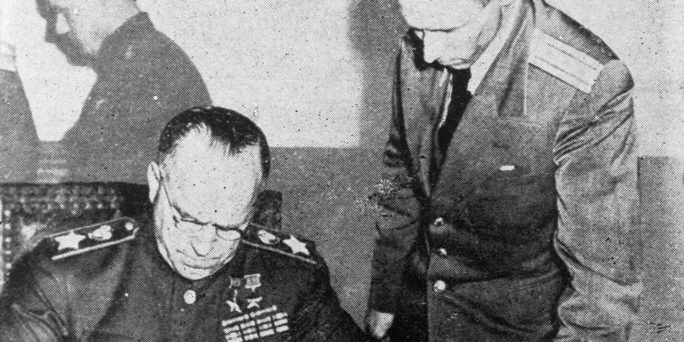 Man seated a table signing a document, with another man standing over him.