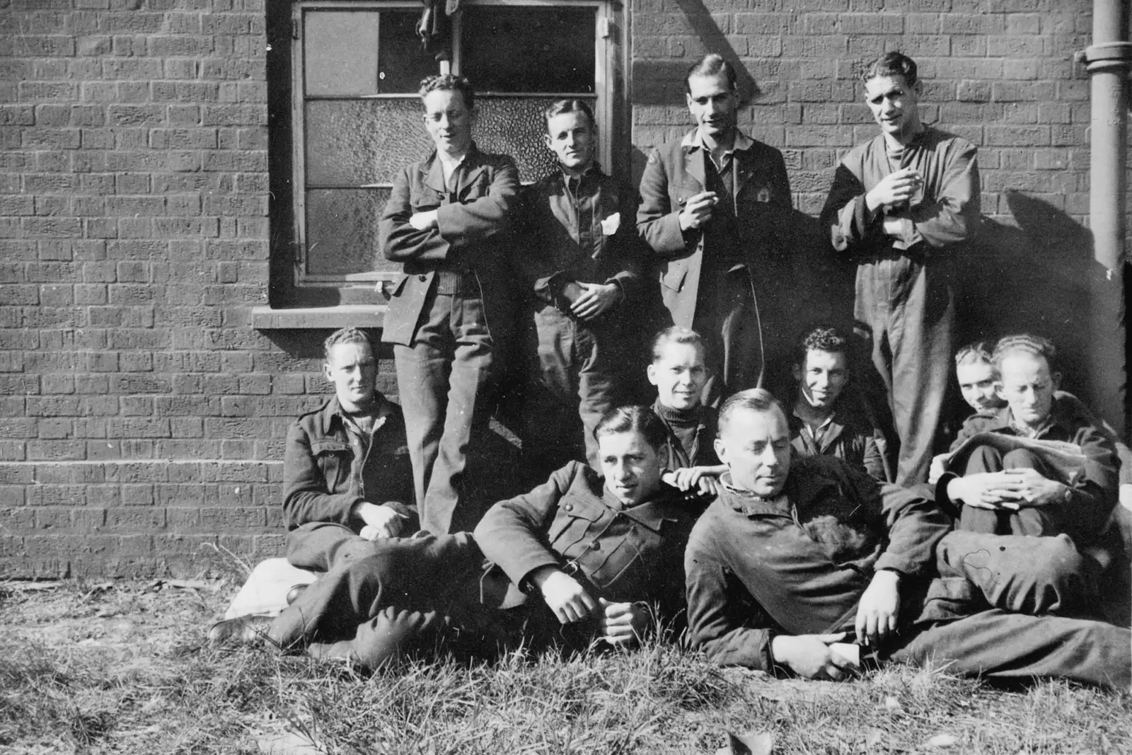 Four men stand against a brick wall, in front are seven men sitting on the ground.