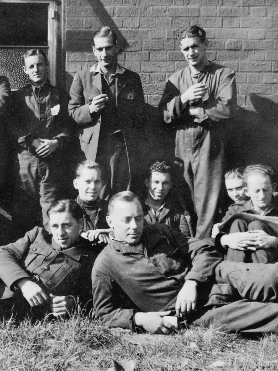 Four men stand against a brick wall, in front are seven men sitting on the ground.