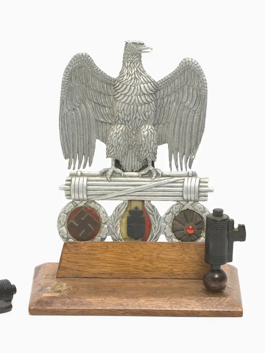  An ornate and fastidiously decorated fascist eagle clasping a Roman fasces, from which are suspended the symbols of the three Axis allies, each contained within a decorated wreath - from left to right, the Nazi swastika; the Italian arms and the Japanese 'rising sun' flag. 