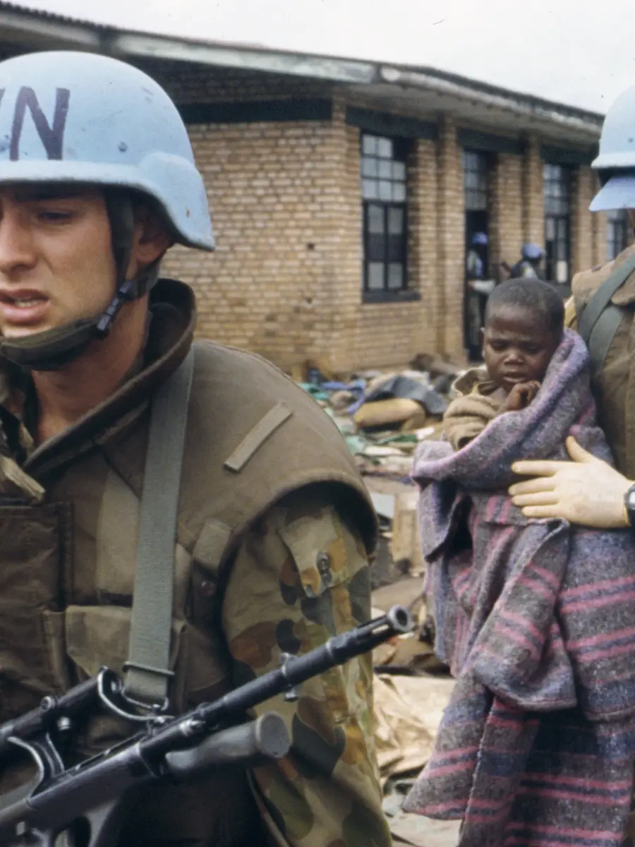 An armed Australian soldier leading another soldier who is carrying a Rwandan child in his arms.