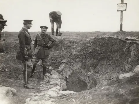 The King of the Belgians and British Army General Sir Hubert Gough at the entrance to a former German Army dugout. An officer in the background is inspecting a German helmet. 
