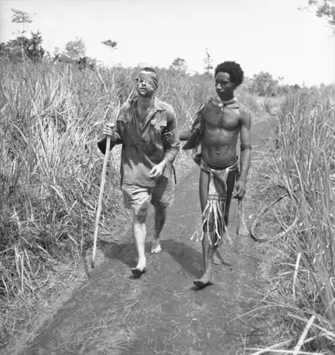 Papuan native Raphael Oimbari helping Private George Whittington towards a hospital.