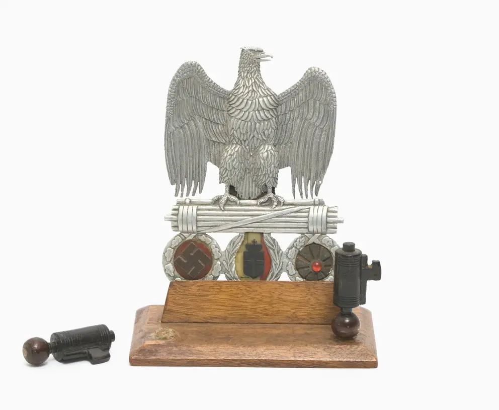  An ornate and fastidiously decorated fascist eagle clasping a Roman fasces, from which are suspended the symbols of the three Axis allies, each contained within a decorated wreath - from left to right, the Nazi swastika; the Italian arms and the Japanese 'rising sun' flag. 