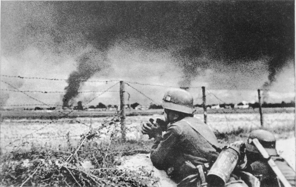Two German troops lay on the ground, looking at smoke in the distance.